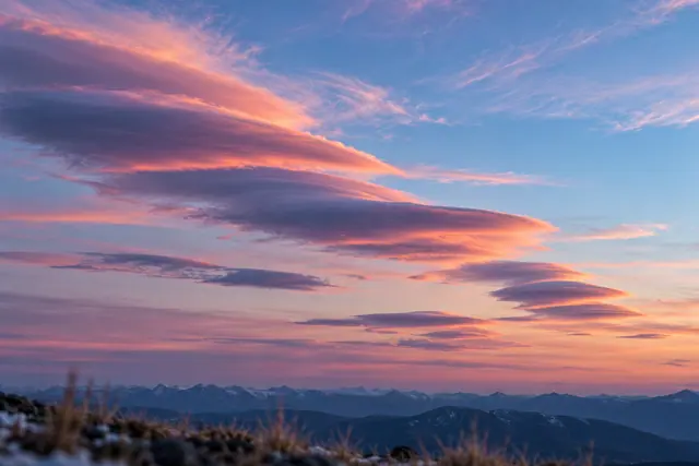 Nuvens lenticulares rosadas e lilases no céu do crepúsculo sobre montanhas, com capim desfocado em primeiro plano.