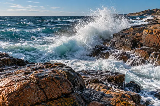 Onda quebra em rochas alaranjadas, espalhando espuma e gotas diante do mar azul e do céu com nuvens finas.