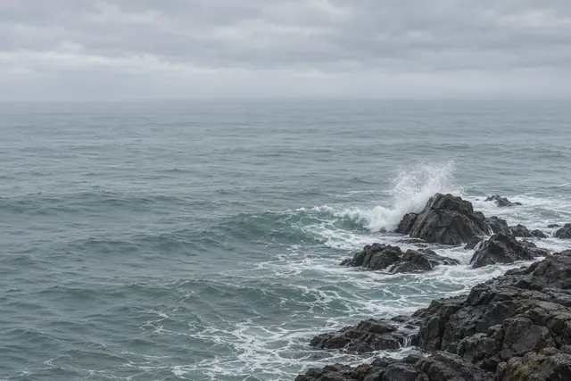 Ondas quebram em rochas escuras à direita, com espuma branca, mar azul-acinzentado e céu nublado ao fundo.