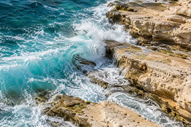Onda turquesa quebra em rochas claras e espalha espuma branca sob a luz do sol, em vista elevada.