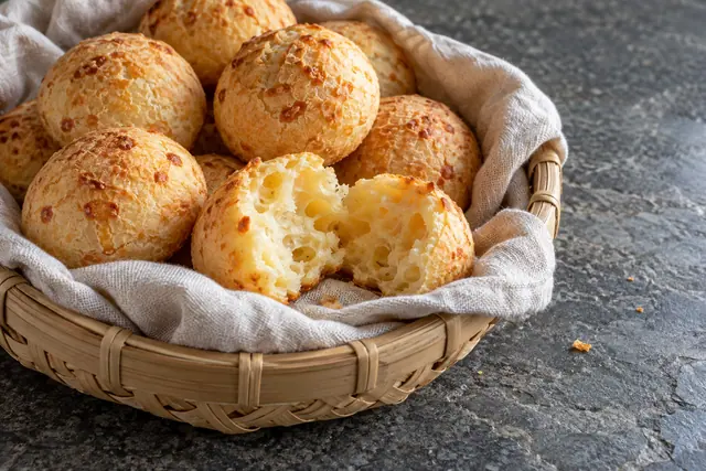 Cesta de bambu com pães de queijo dourados e um aberto ao meio sobre tecido claro e bancada de pedra cinza.