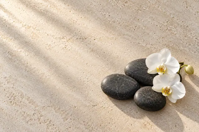Três pedras de spa com orquídeas brancas sobre pedra clara, sob luz suave e sombras diagonais.