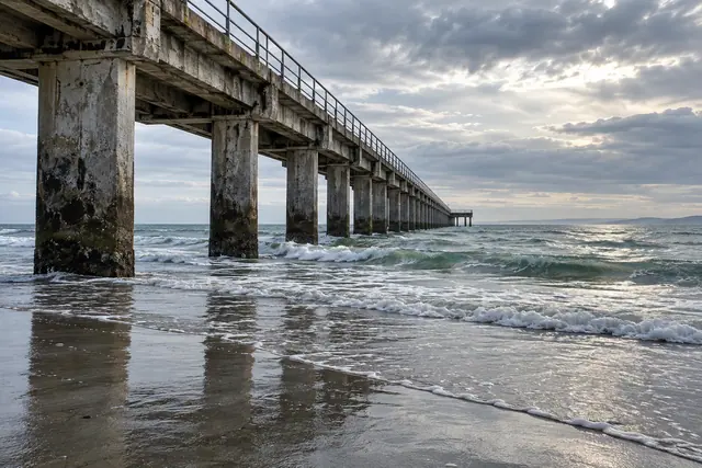 Píer de concreto visto de lado, com ondas, reflexos na areia molhada e céu nublado sobre o mar.
