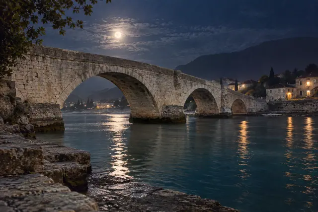 Ponte de pedra com três arcos sob o luar, com reflexos no rio, casas iluminadas e montanhas ao fundo.