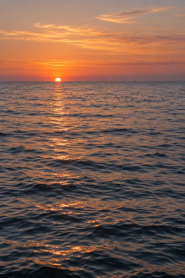 Sol no horizonte sobre o mar calmo, com reflexos dourados nas ondas e nuvens em um céu alaranjado.
