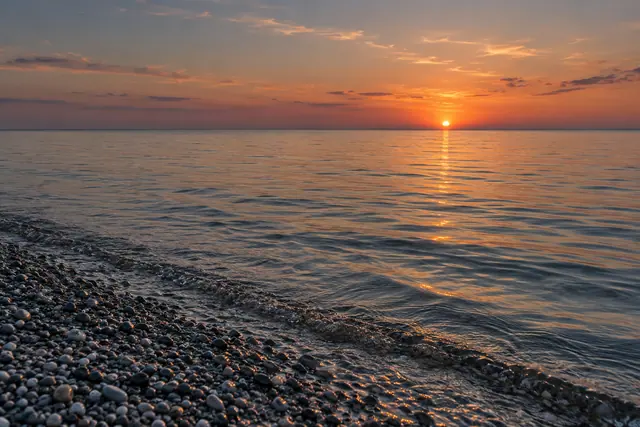 Pôr do sol alaranjado sobre o mar calmo, com reflexo dourado na água e seixos molhados na margem.
