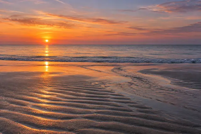 Pôr do sol alaranjado sobre mar tranquilo, com reflexos dourados na areia ondulada e um canal de água na praia.