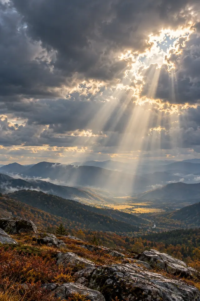 Raios de sol atravessam nuvens escuras sobre um vale montanhoso com névoa, vegetação de outono e pedras.