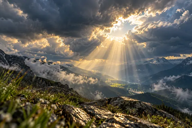 Raios de luz atravessam nuvens escuras sobre um vale verde cercado por montanhas e névoa.