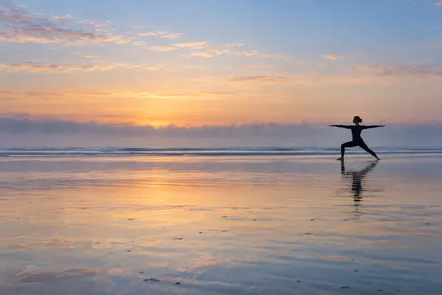 Pessoa em silhueta pratica yoga na areia molhada, refletida diante do mar sob o céu azul e pêssego do amanhecer.