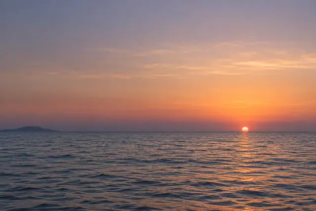 Sol se põe sobre o mar calmo, com reflexo dourado, céu em tons suaves e uma ilha distante à esquerda.
