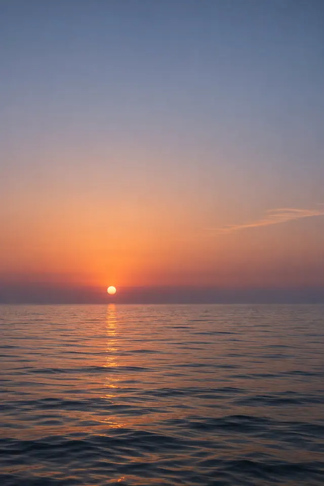 Sol baixo sobre o mar calmo, com reflexo dourado e céu amplo em tons alaranjados e azul acinzentado.