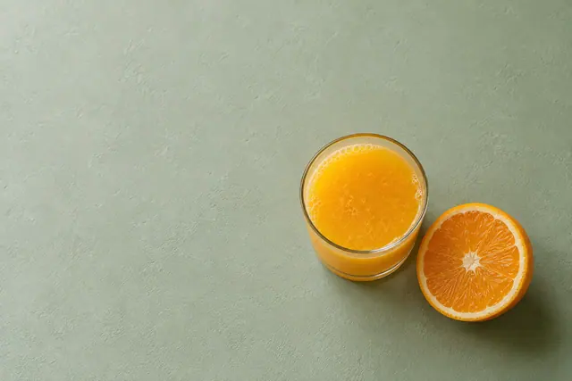 Copo de suco de laranja e meia laranja sobre fundo verde-sálvia, com luz suave e amplo espaço livre.