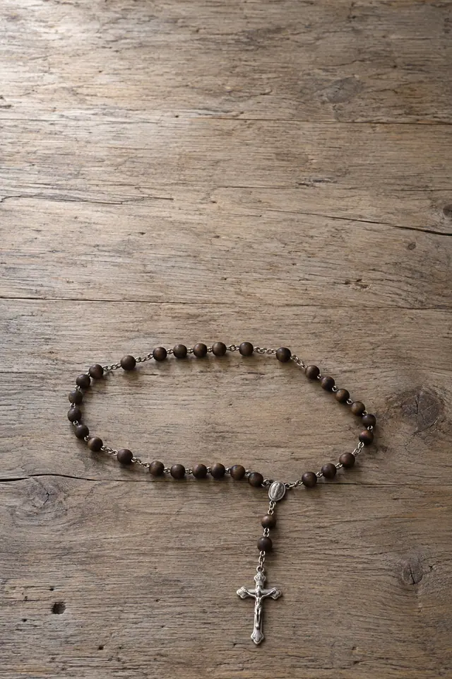 Terço de contas escuras e crucifixo prateado sobre mesa de madeira antiga, com luz suave e amplo espaço livre.