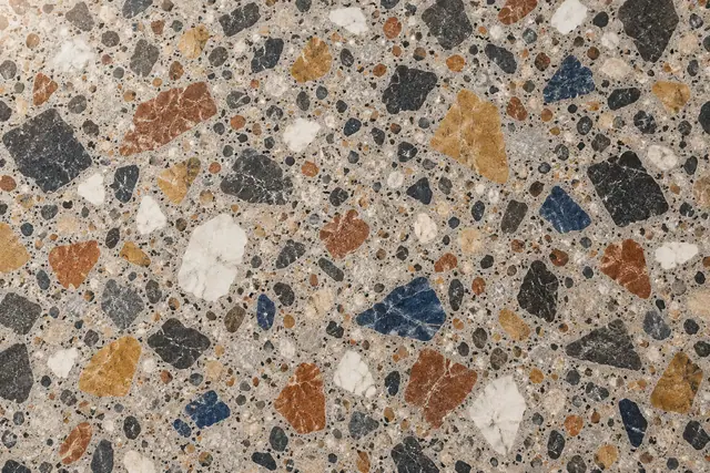 Textura de terrazzo cinza-claro com fragmentos irregulares em preto, terracota, ocre, branco e azul-profundo.