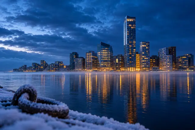 Prédios iluminados refletem na água sob céu azul nublado, com cais nevado e argola desfocada em primeiro plano.