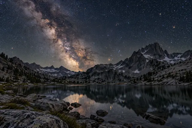Via Láctea sobre montanhas rochosas com neve, refletida em um lago com pedras na margem.