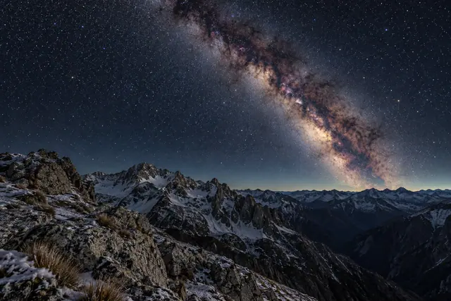 Via Láctea em diagonal sobre montanhas nevadas, com rochas e capim em primeiro plano e céu repleto de estrelas.