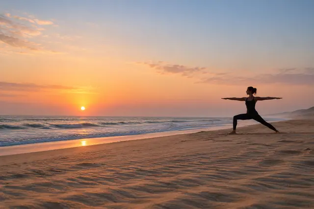 Mulher pratica yoga na areia da praia ao amanhecer, com braços estendidos, ondas suaves e céu em tons de pêssego.